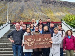 Zdjęcie przedstawia grupę 15 osób, która stoi przed budynkiem i trzyma tablicę z napisem FAB LAB Ísafjörður. 